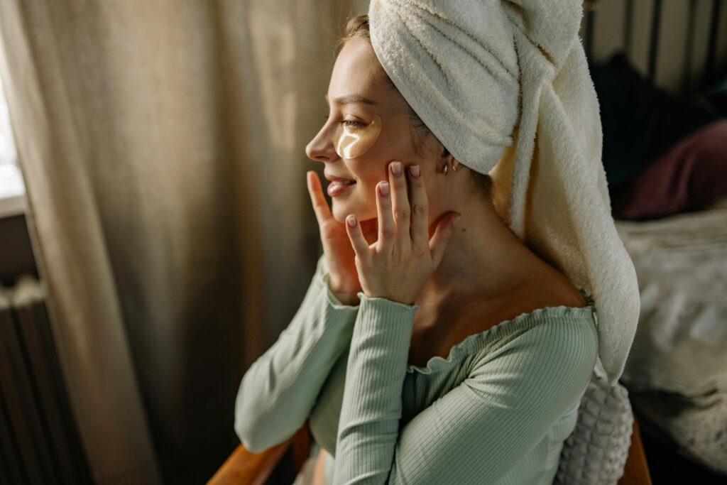 A woman enjoying a skincare routine with eye patches and a towel wrapped on her head indoors.